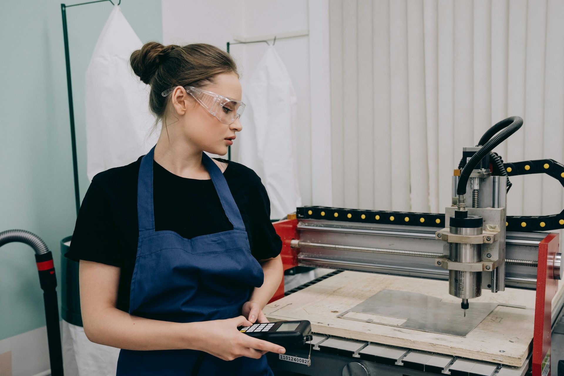 Mujer en taller utilizando máquina CNC, con gafas protectoras y delantal, centrada en el trabajo industrial.